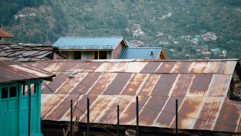Rustic metal-roofed houses in Manali, nestled against lush green mountains under a blue sky.