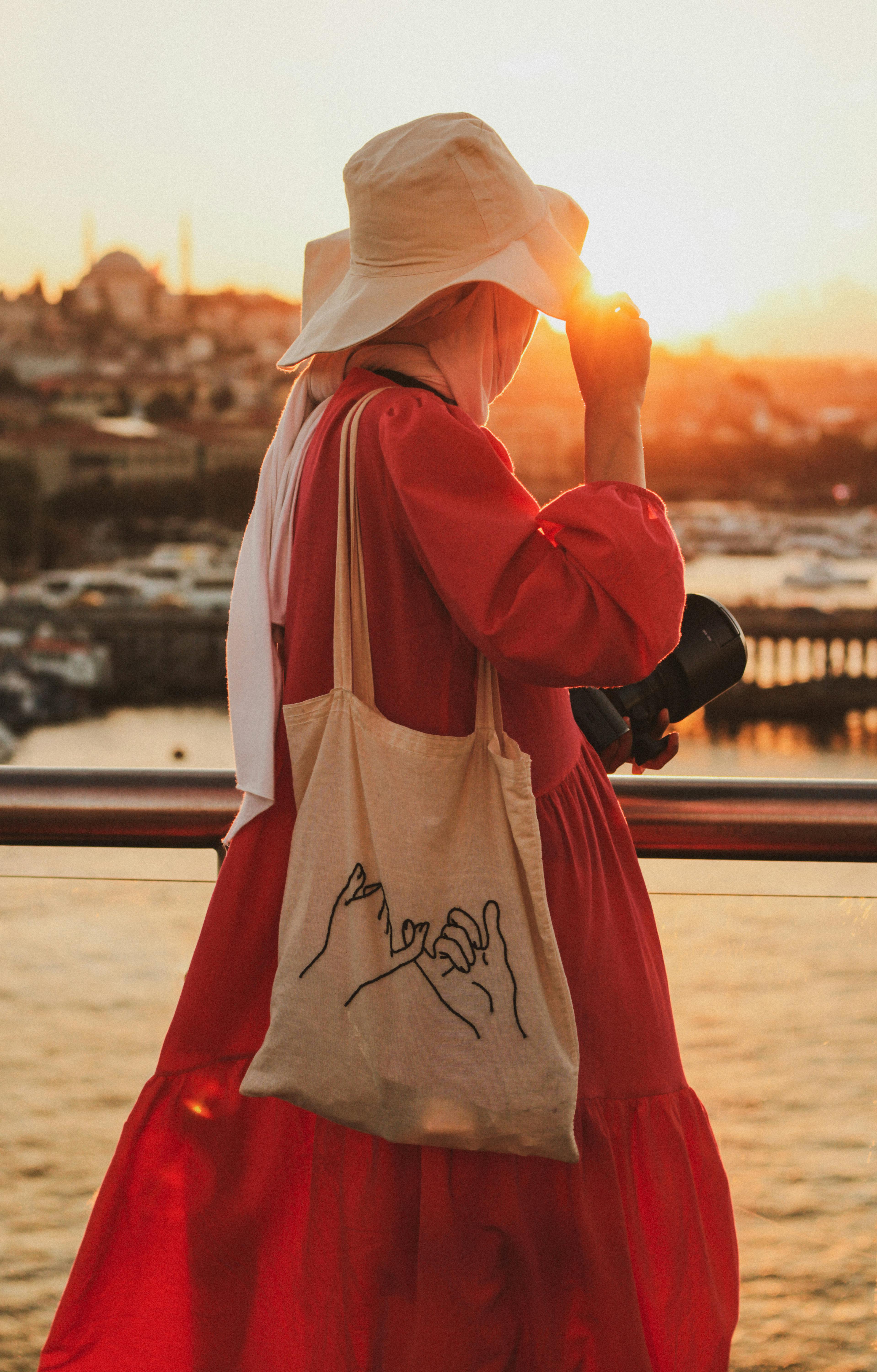 Woman in a bucket hat on a balcony at sunset, holding a camera and wearing an orange dress.