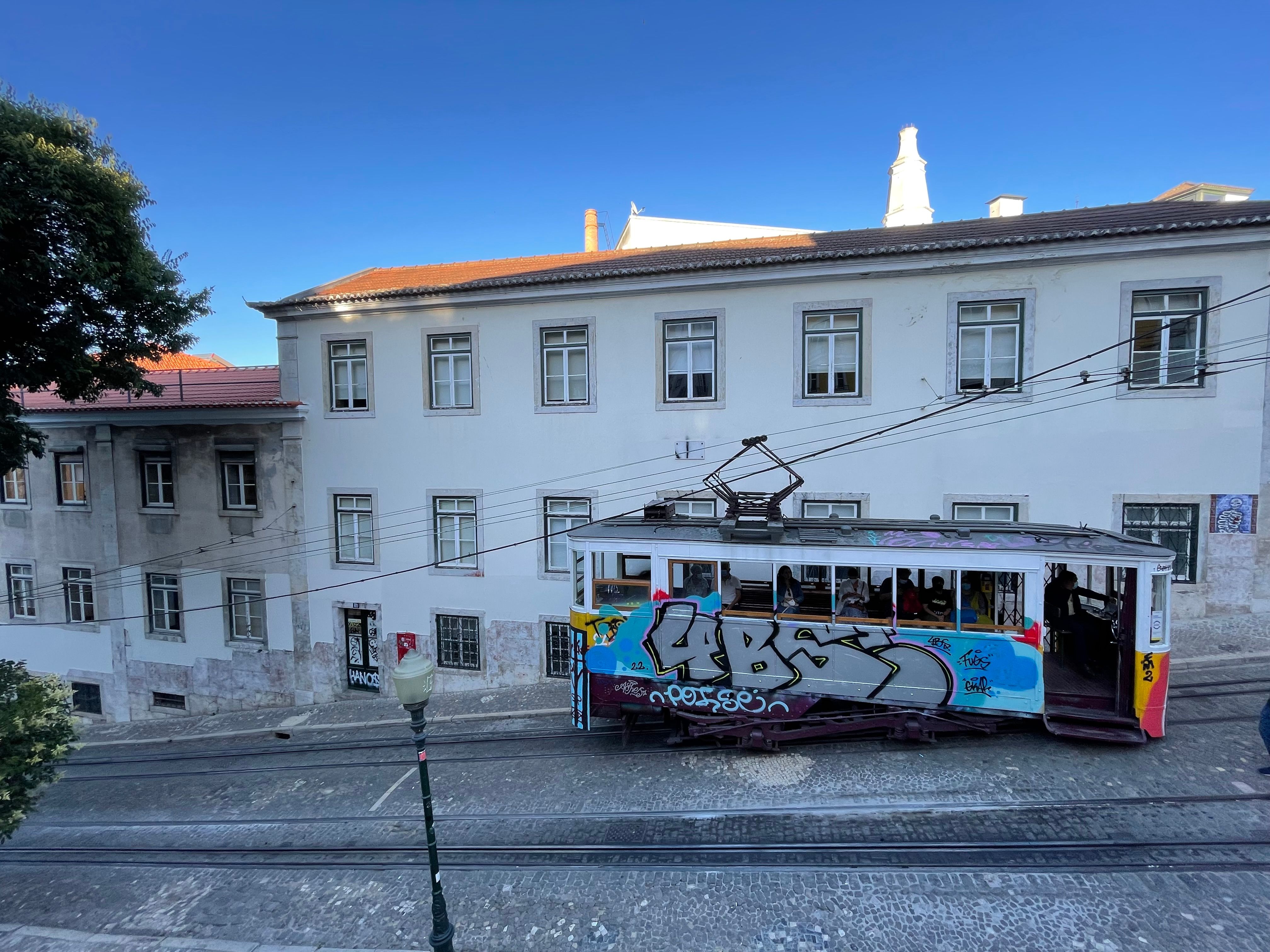 Historic yellow tram winding through a narrow, hilly street in Lisbon's Alfama district, with colorful buildings and laundry hanging outside windows.