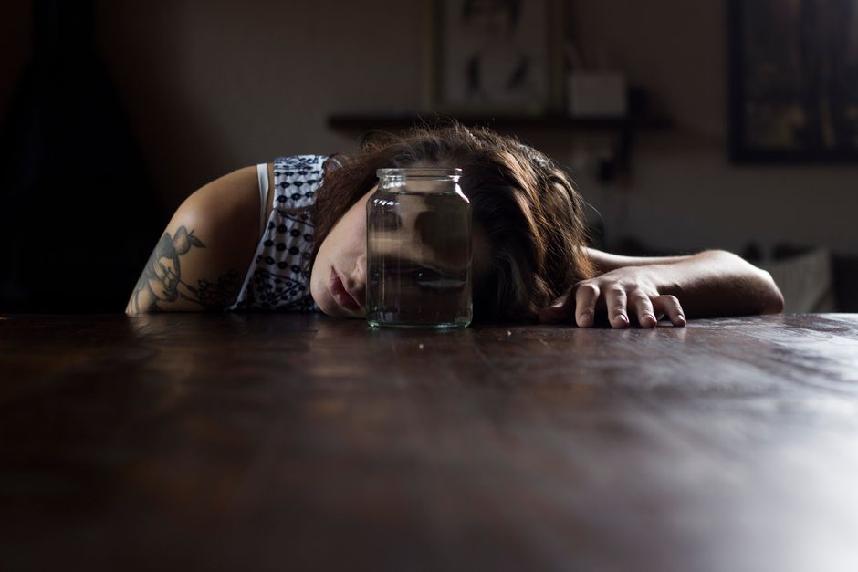 A pensive woman with tattoos leans on a table next to a mason jar in a dimly lit room.
