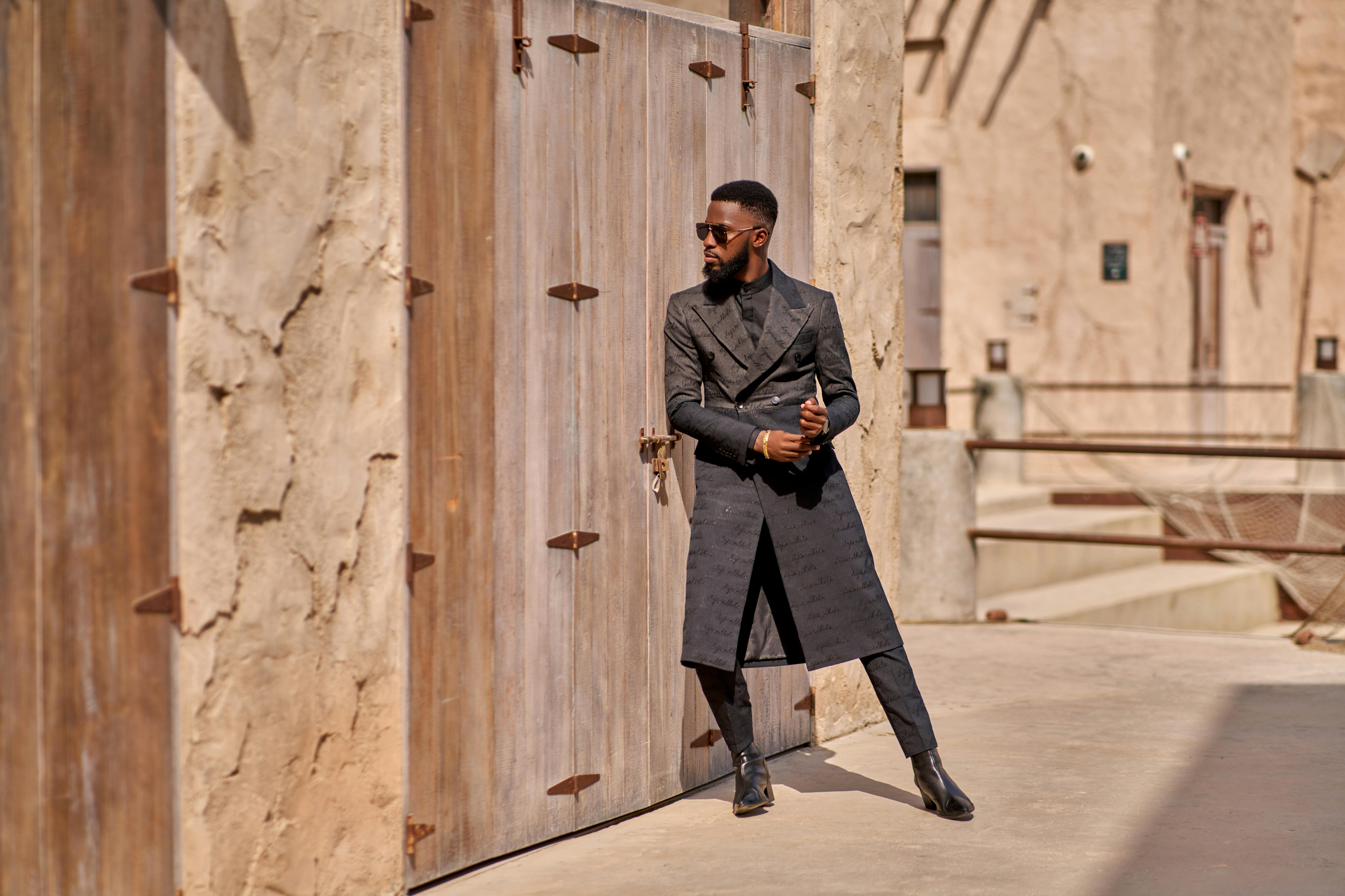 Fashion-forward man in Dubai wearing a long coat and sunglasses, leaning on rustic doors.