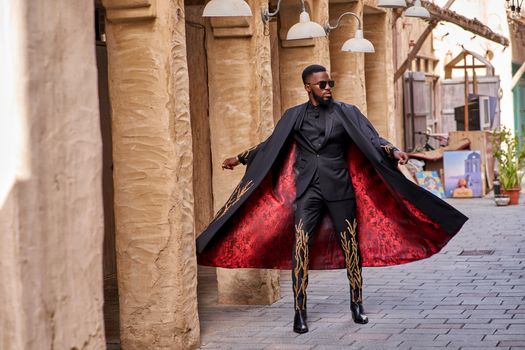 Fashion-forward man in a trendy black suit with red interior posing outdoors against rustic columns.