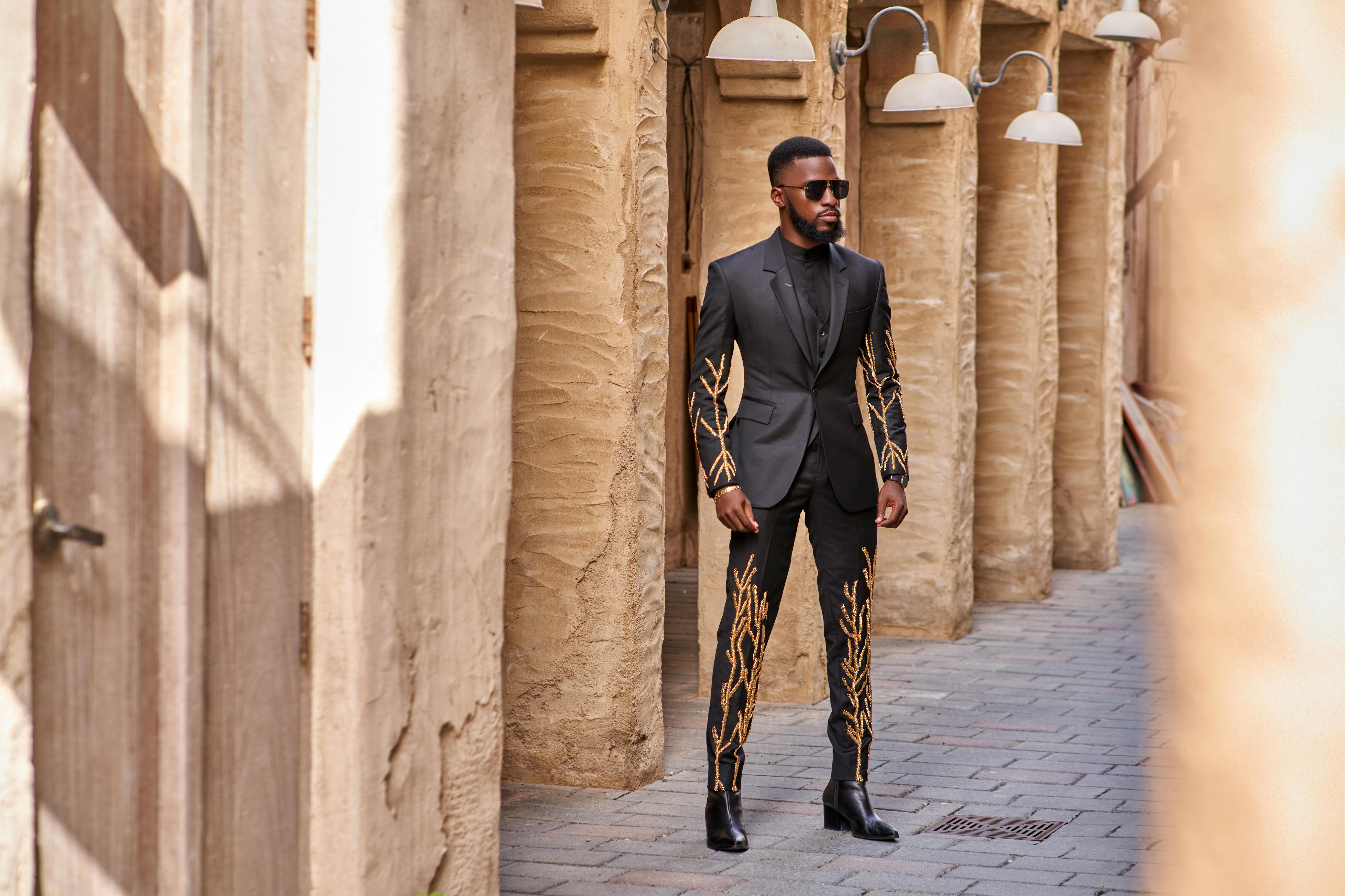 A fashionable man in an embroidered suit stands confidently in a rustic alleyway. Urban elegance meets tradition.