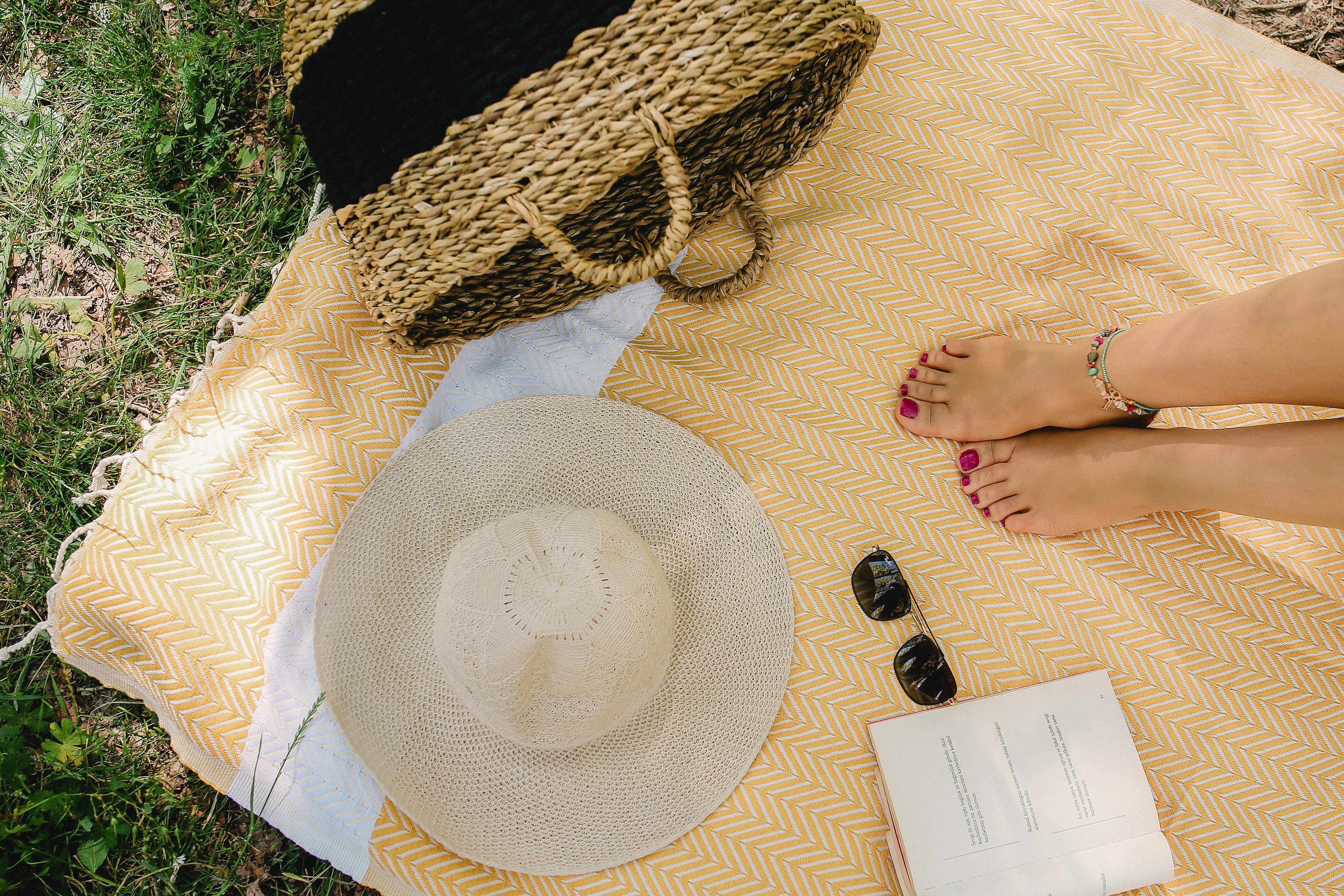 A serene picnic setup with feet, straw hat, sunglasses, and a woven bag under summer sun.