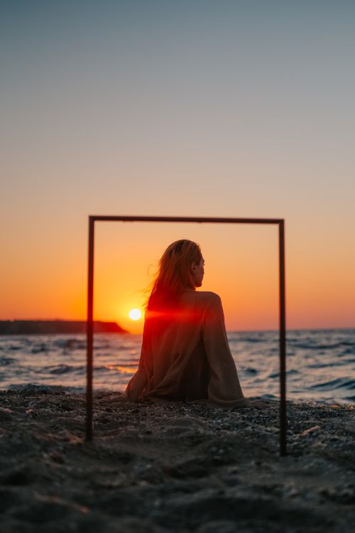 Free Back view of a woman sitting on a sandy beach at sunset, framed by a minimal structure, creating a peaceful scene. Stock Photo