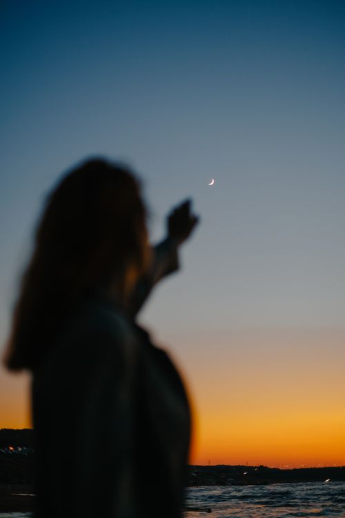 Free Silhouette of a woman pointing at the crescent moon against a vibrant sunset sky. Stock Photo