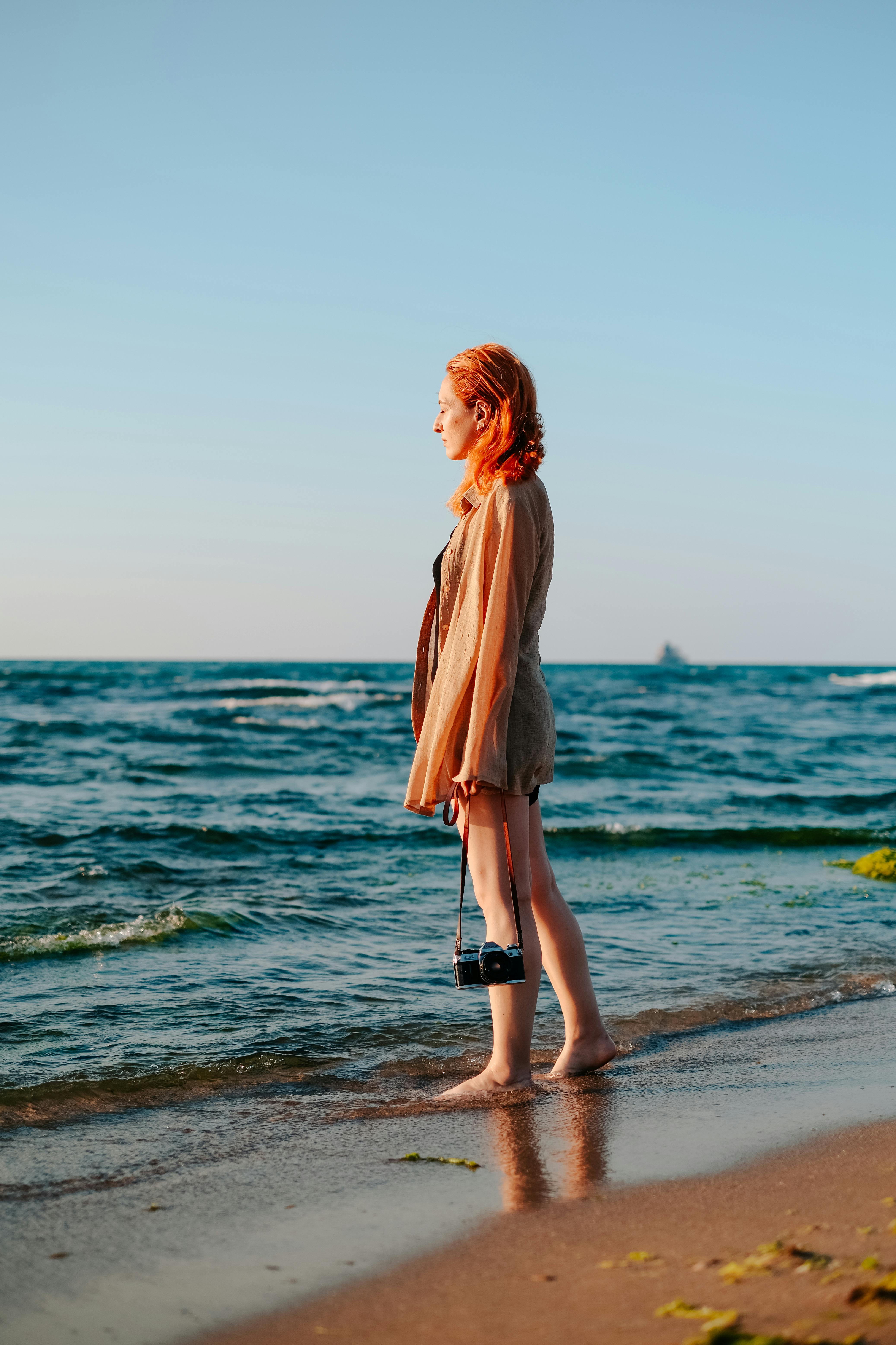 A woman stands on a beach during sunset, holding a camera and enjoying the ocean view.