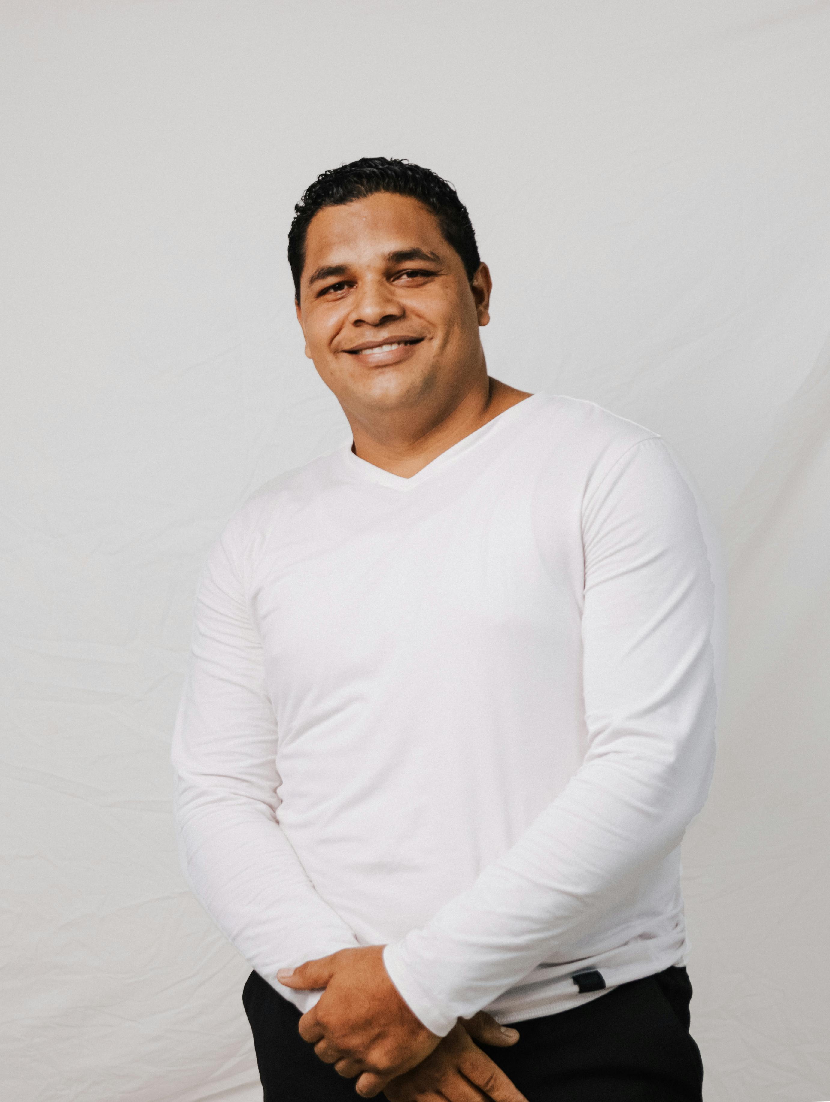 Studio portrait of a smiling adult man in a white shirt standing against a white background.