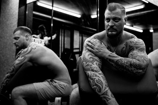 Black and white portrait of a tattooed man in a gym, reflecting in mirrors.