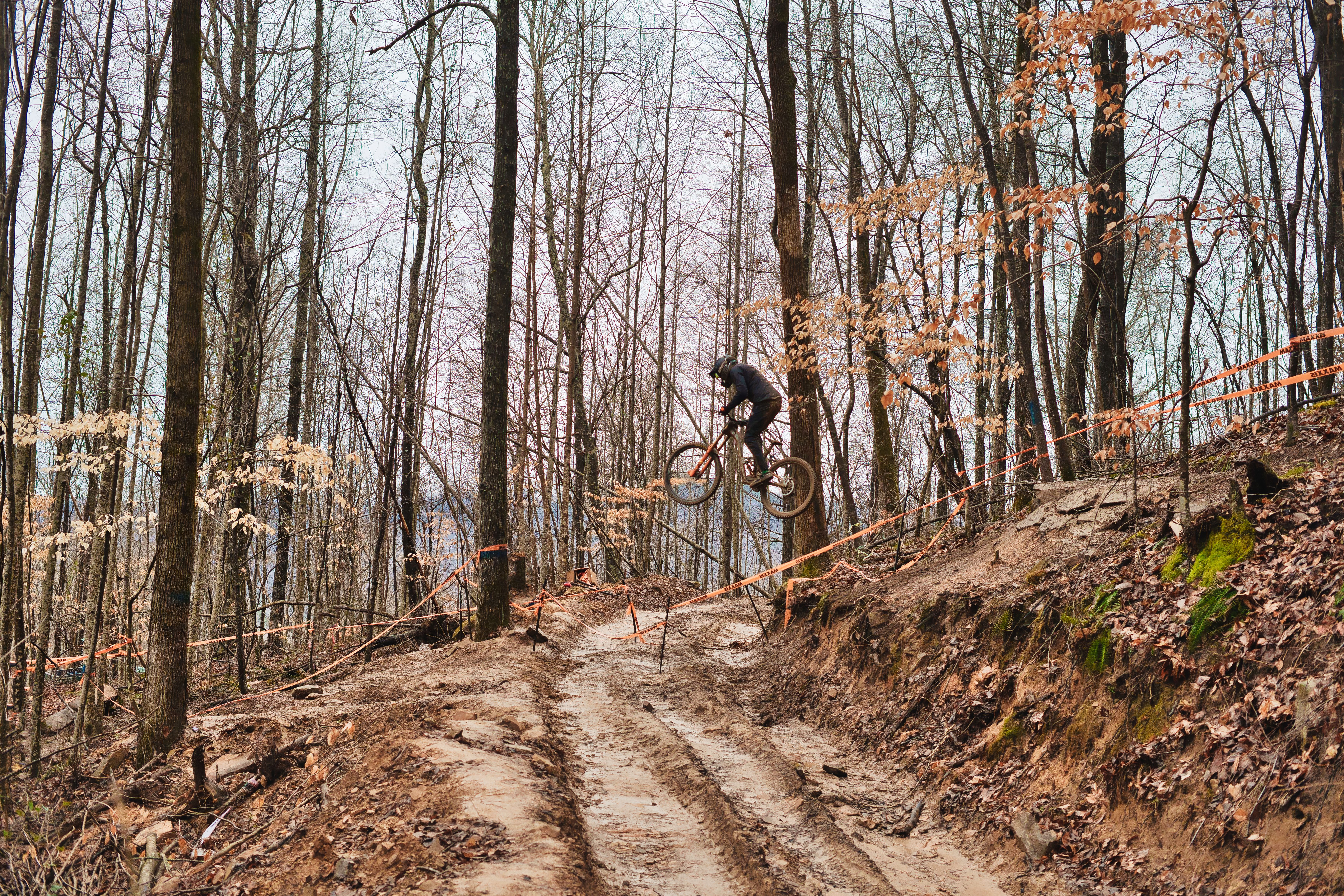 Rider mid-air on a full-suspension enduro mountain bike over a muddy forest trail marked with Maxxis race tape.