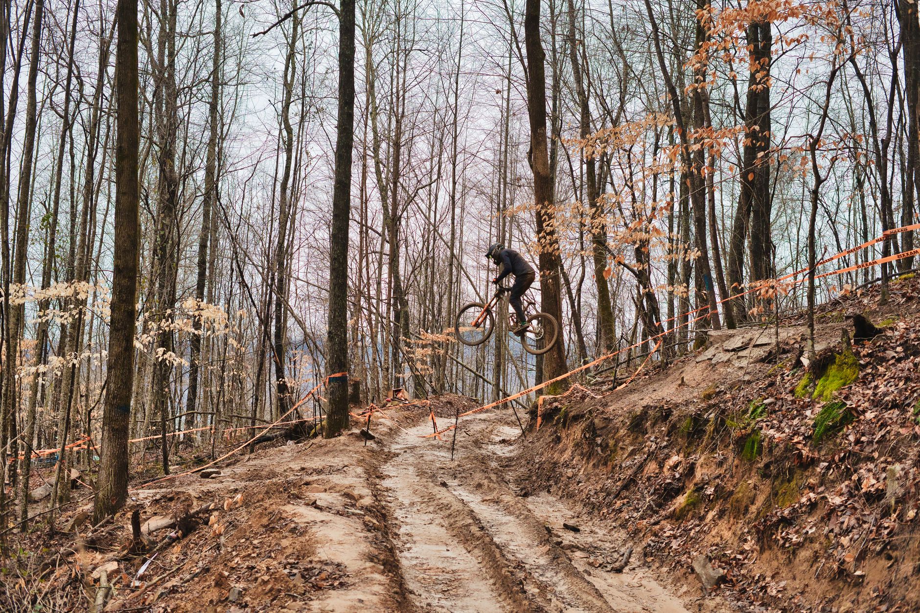 Rider mid-air on a full-suspension enduro mountain bike over a muddy forest trail marked with Maxxis race tape.