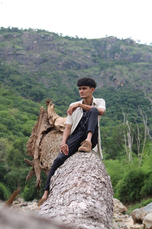 Free Young man sitting on a fallen tree in a scenic mountain setting. Perfect for outdoor and adventure themes. Stock Photo