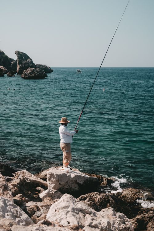 Free A man enjoys fishing on a rocky seaside with a clear ocean view. Stock Photo