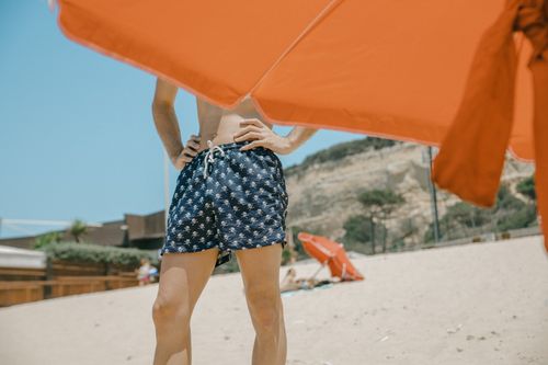 Free Man in patterned swim shorts enjoying a sunny day under an orange beach umbrella. Stock Photo
