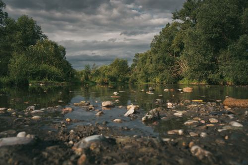 Free Scenic view of a peaceful river with rocks and lush greenery beneath a moody cloudy sky. Stock Photo