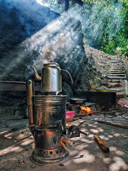 Sunbeams illuminate a vintage metal kettle over an outdoor fire beside a rustic stone wall.