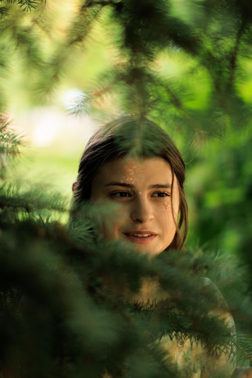 Free A young woman smiles subtly among lush evergreen branches, conveying a serene and natural vibe. Stock Photo
