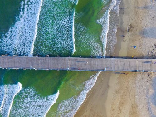 Free A stunning aerial shot of a pier stretching over crashing ocean waves, with a sandy beach beside it. Stock Photo