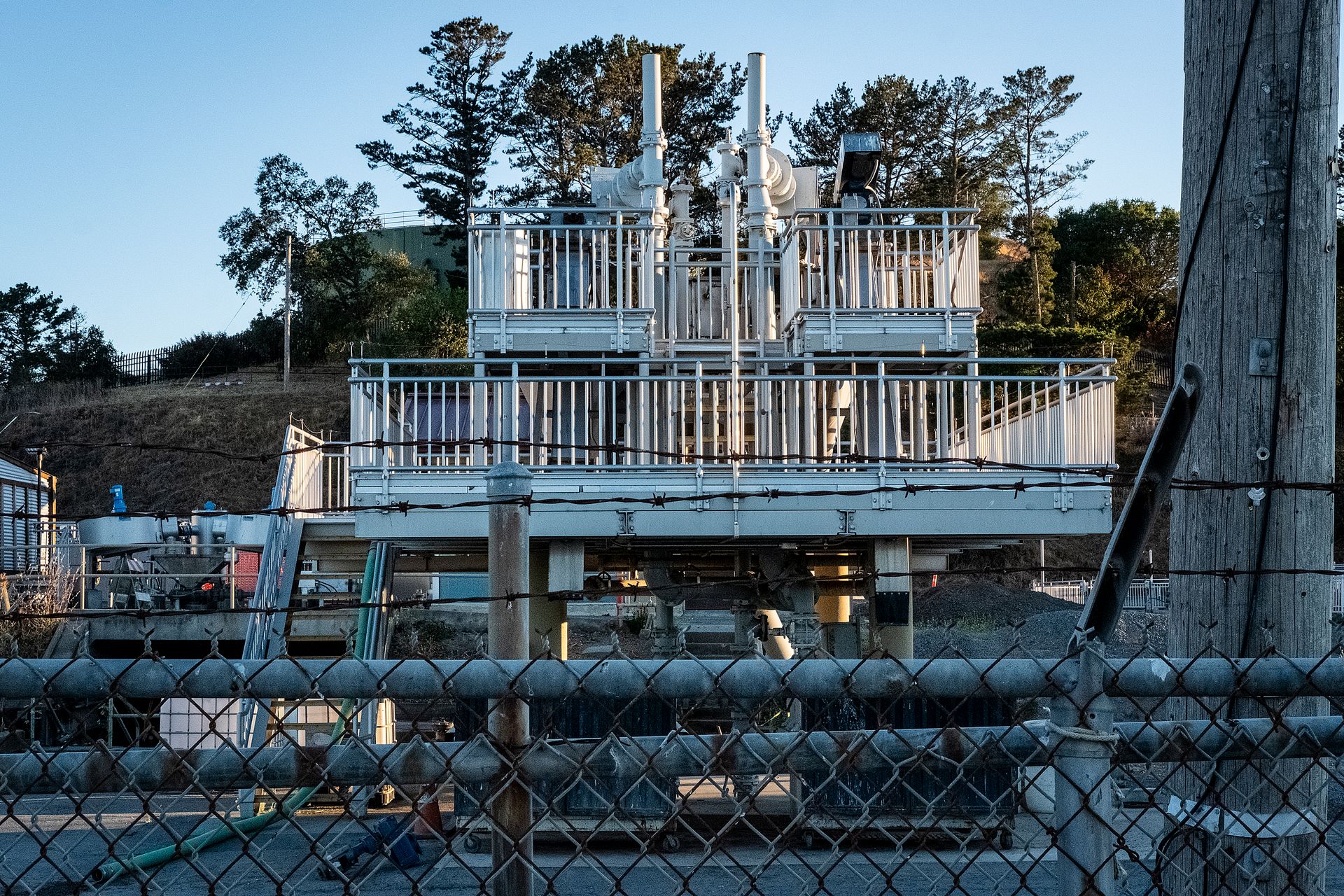 An outdoor industrial facility featuring metal structures and barbed wire fence, photographed at sunset.