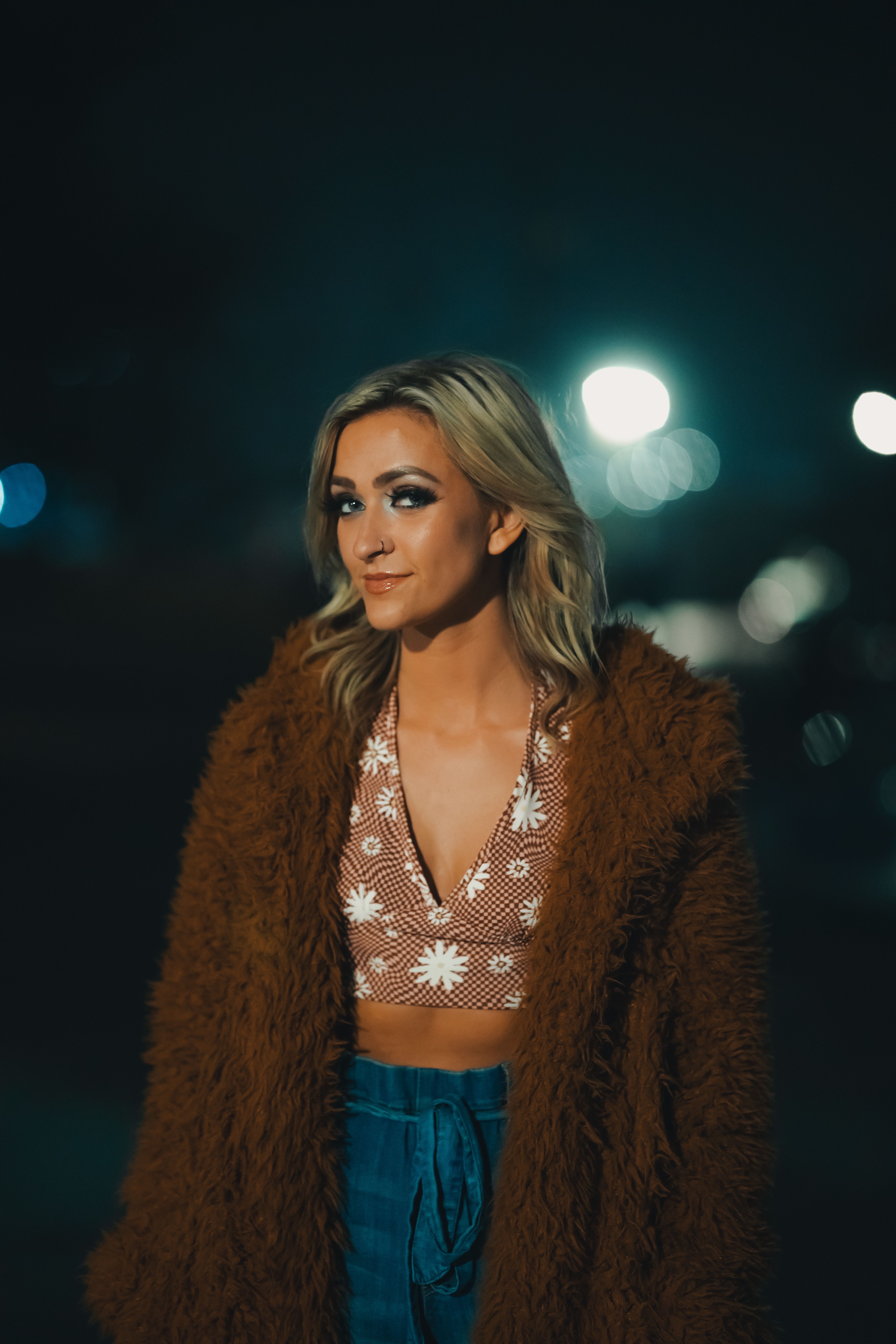 A fashionable woman stands outdoors at night wearing a fur coat with glowing lights in the background.