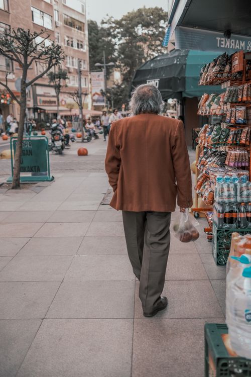 Free Senior man in brown coat walking past city street market stalls on a busy sidewalk. Stock Photo