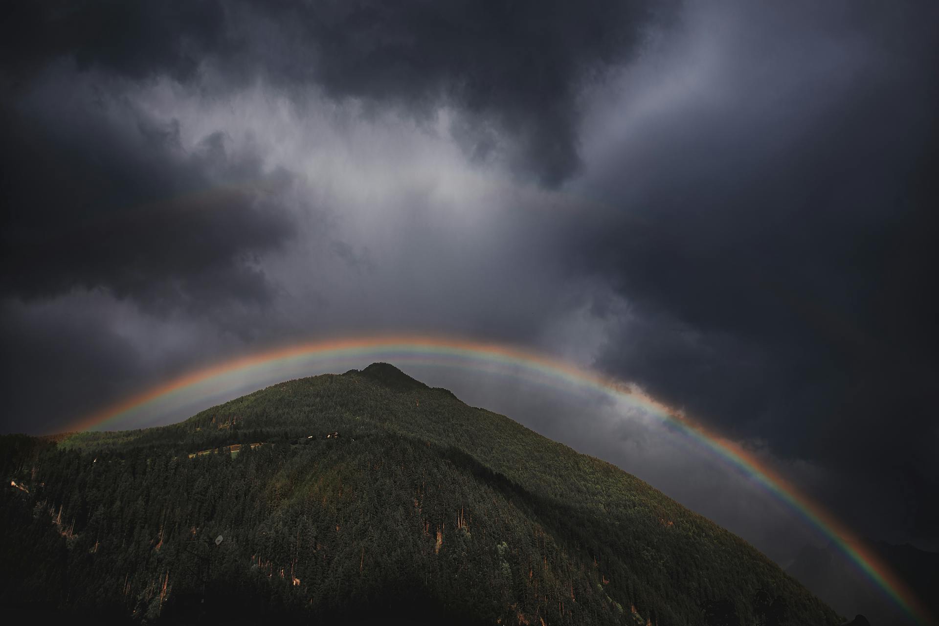 Dark storm clouds over a lush mountain with a vibrant rainbow arching across the sky.