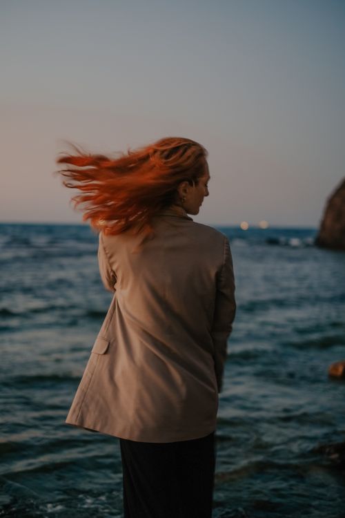Free A woman stands near the sea with her hair flowing in the wind, capturing a serene and dynamic moment. Stock Photo