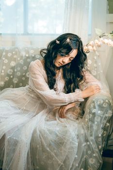 Beautiful woman in a white dress with flowers in her hair sitting indoors, exuding elegance and grace.