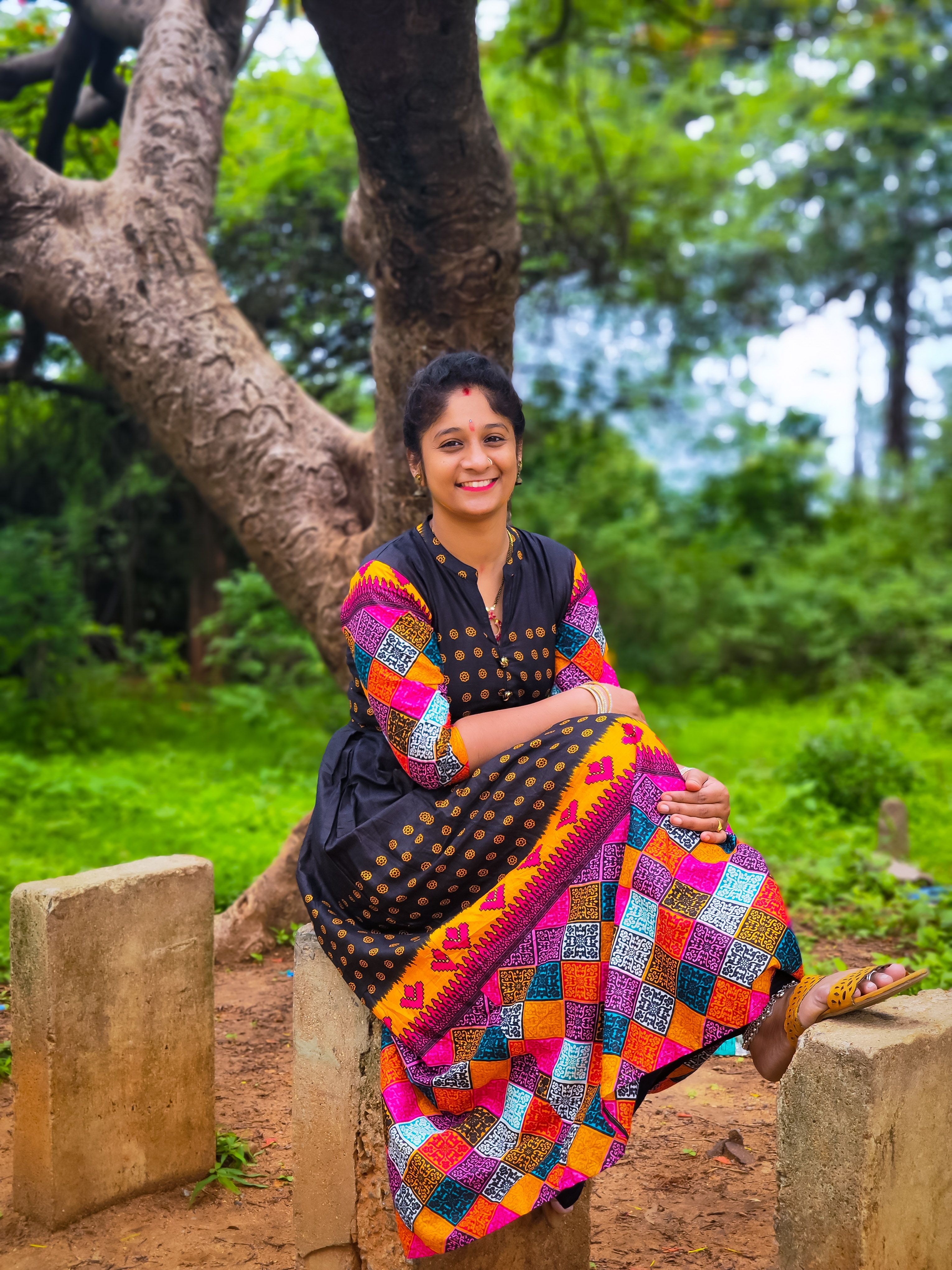 South Asian woman in vibrant dress seated outdoors on a sunny day.