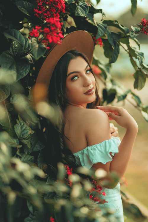 Free Brunette woman in a mint dress and hat surrounded by lush greenery and red flowers, exuding elegance and style. Stock Photo
