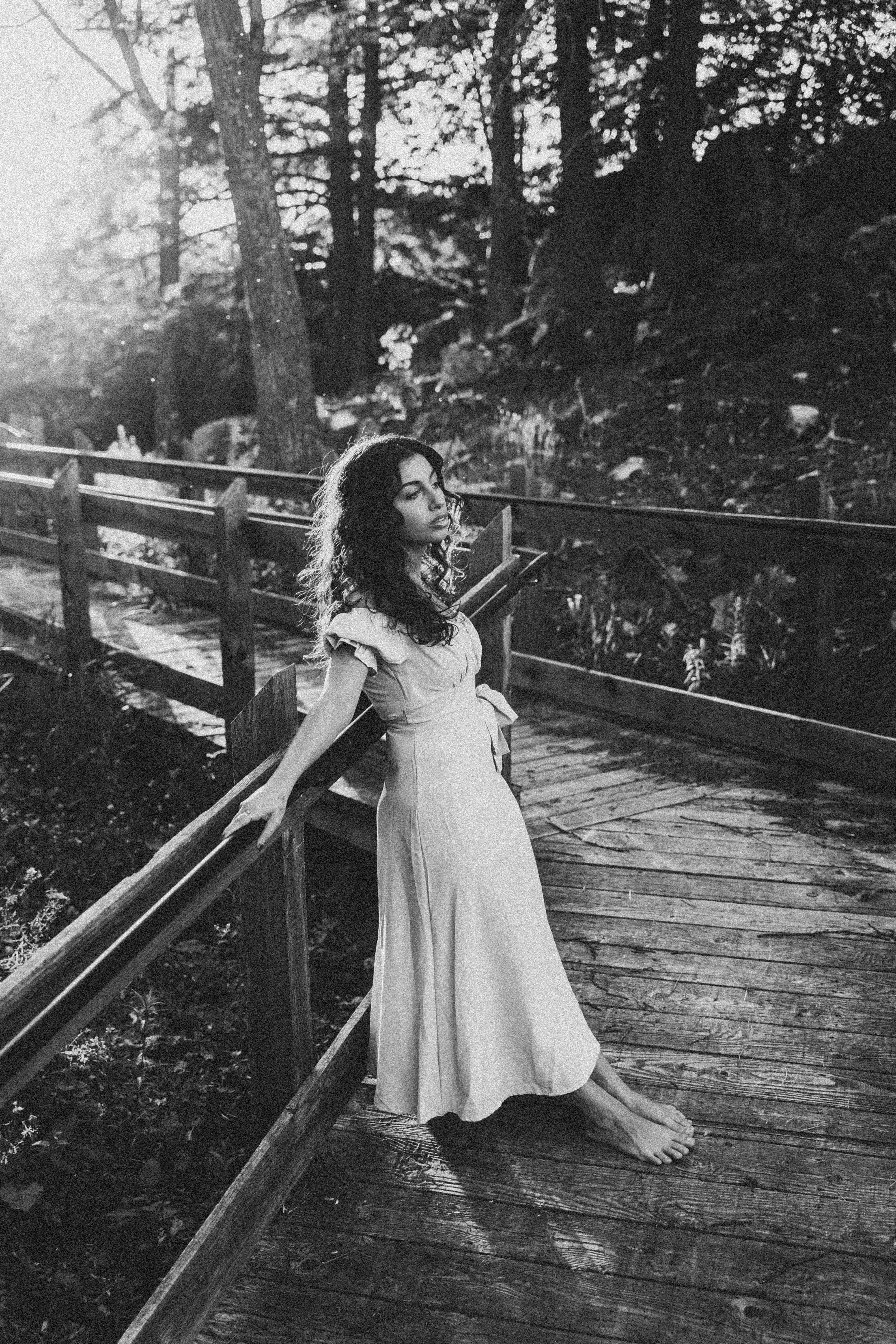 Black and white photo of a woman in a dress posing on a wooden bridge in a serene forest setting.