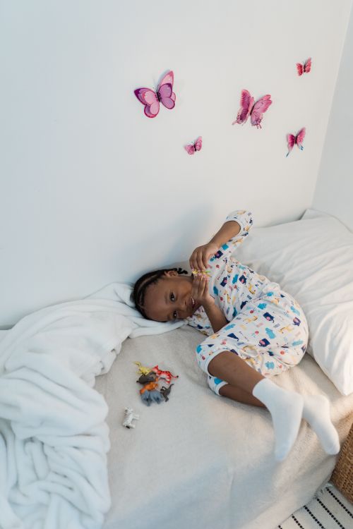 Free Child in pajamas lying in bed surrounded by toys and butterfly wall decor in a cozy bedroom. Stock Photo