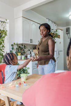 A mother and her daughter share a playful moment at the kitchen table surrounded by toys and greenery.