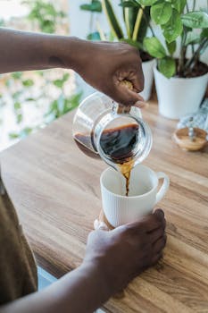 Close-up of hands pouring black coffee from glass pot into mug on a wooden kitchen counter.