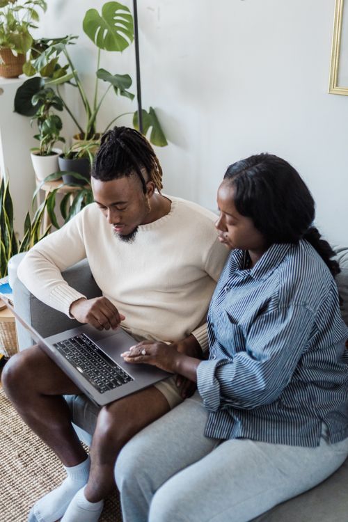 Free A couple sits on a sofa using a laptop in a cozy living room surrounded by indoor plants. Stock Photo