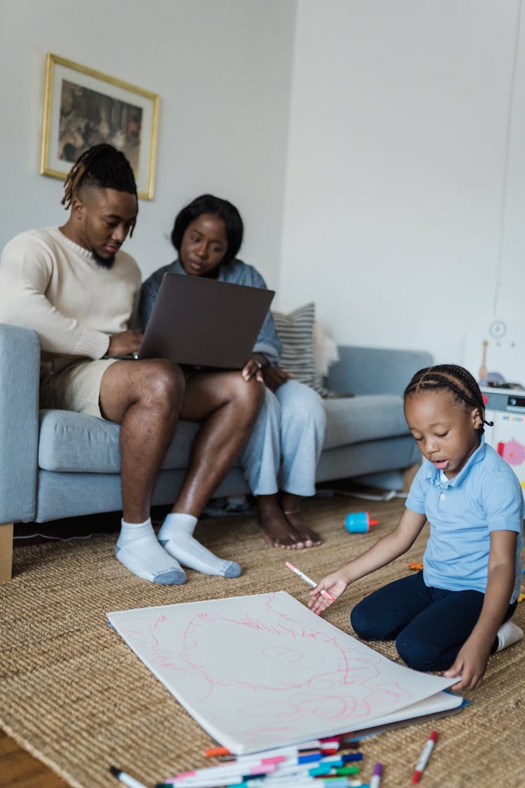 Little Boy Drawing And Sitting On The Floor And Parents Using A Laptop While Sitting On A Couch 