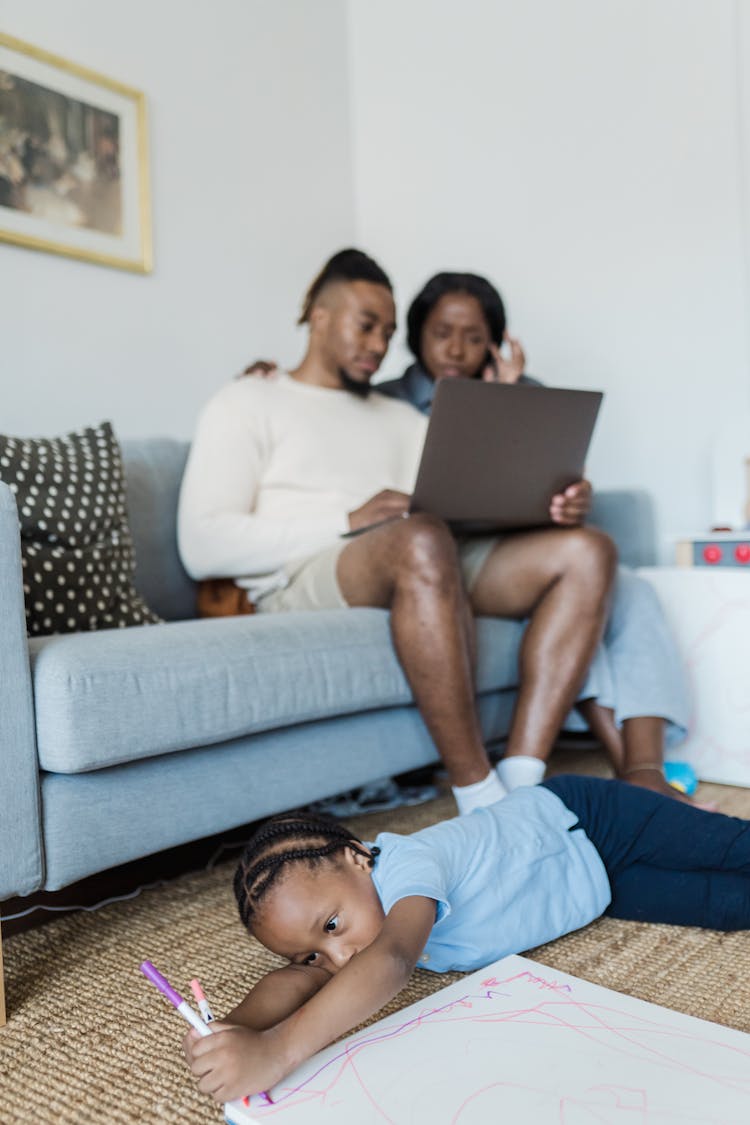 Little Boy Drawing And Lying On The Floor And Parents Using A Laptop While Sitting On A Couch 