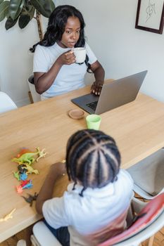 A mother works remotely from home, sipping coffee, as her child plays with toy dinosaurs nearby.