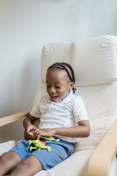 Young boy smiling while playing with a toy dinosaur on a chair indoors.