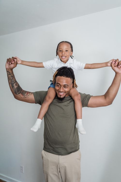 Free Father holding son on shoulders, smiling indoors. Happy family moment. Stock Photo