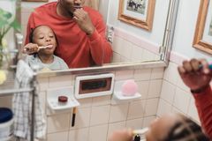 A joyful father and child brushing teeth together in a home bathroom mirror reflection.