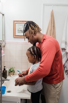 A father helps his daughter brush her teeth in a cozy bathroom setting.