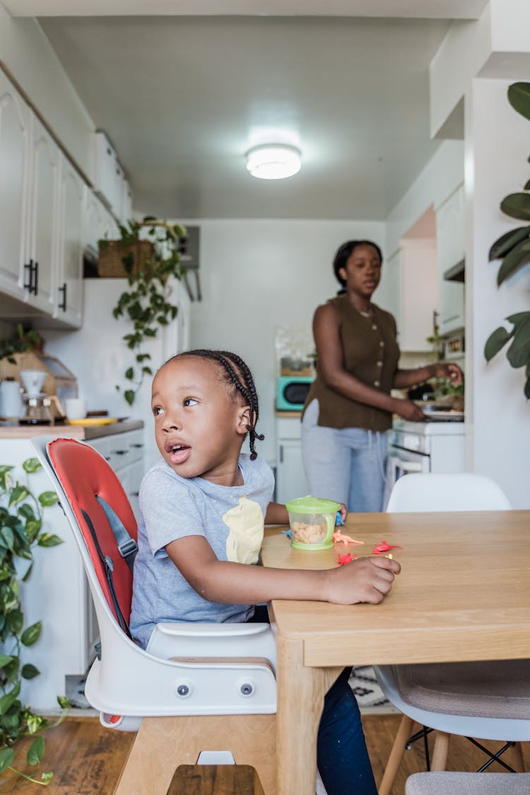 Child Playing With Toys On The Kitchen Table With Mother In The Background 