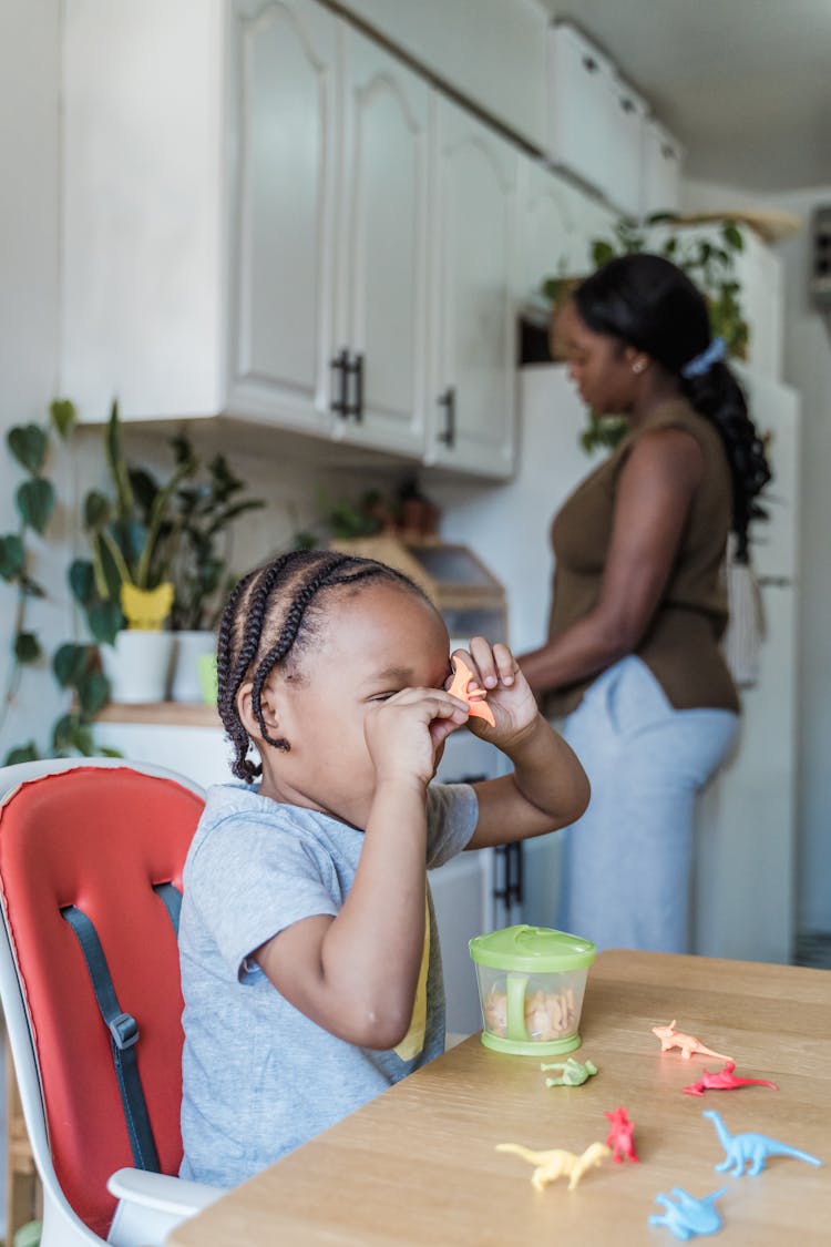 Child Playing With Toys On Kitchen Table With Mother Preparing Food 