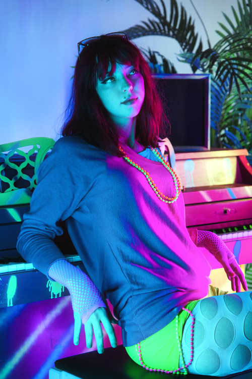 Free A stylish adult woman sits by a piano, illuminated by vibrant neon lighting, creating a modern and artistic ambiance. Stock Photo