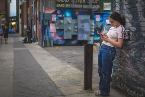 Free Woman in denim and white shirt texting against graffiti wall on an urban street. Stock Photo