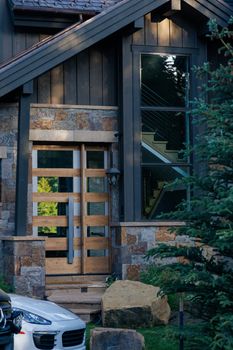 Entrance of a modern rustic house with wooden door and glass window featuring outdoor green plants.