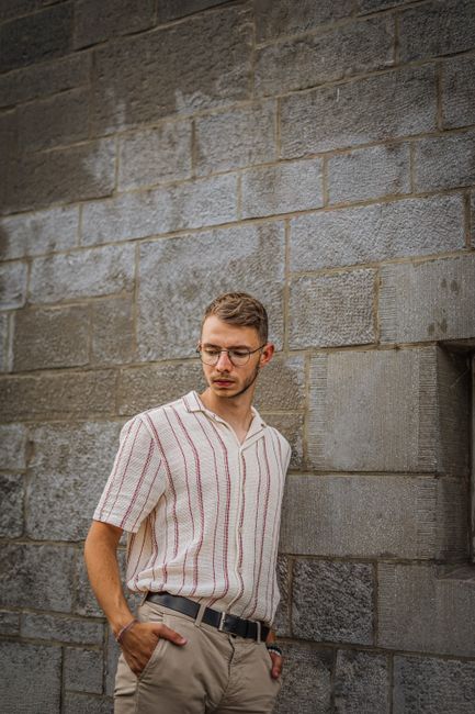 A young man with glasses leans against a stone wall, looking contemplative outdoors.