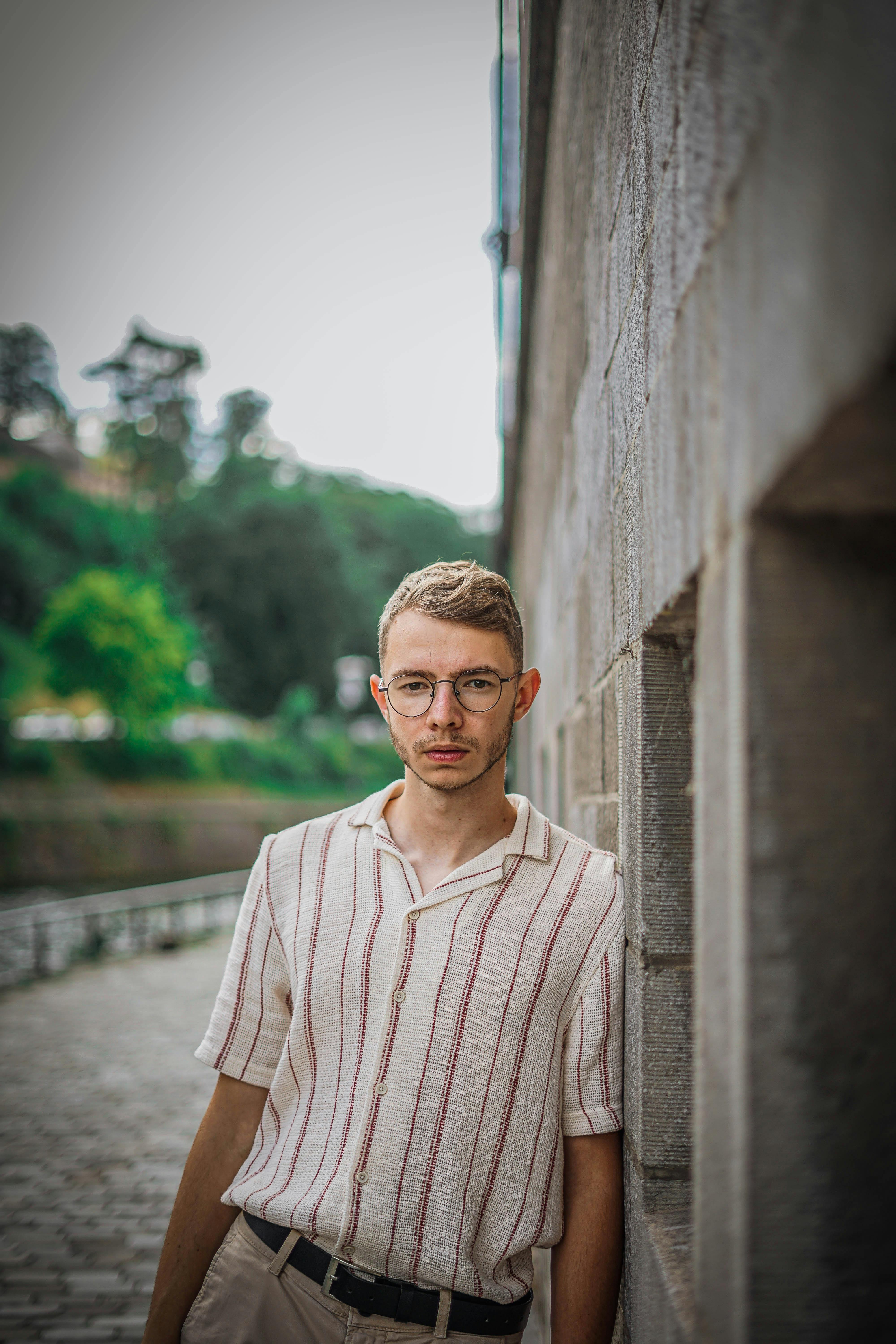 A young man with eyeglasses leans against a textured wall in Namur, Belgium.