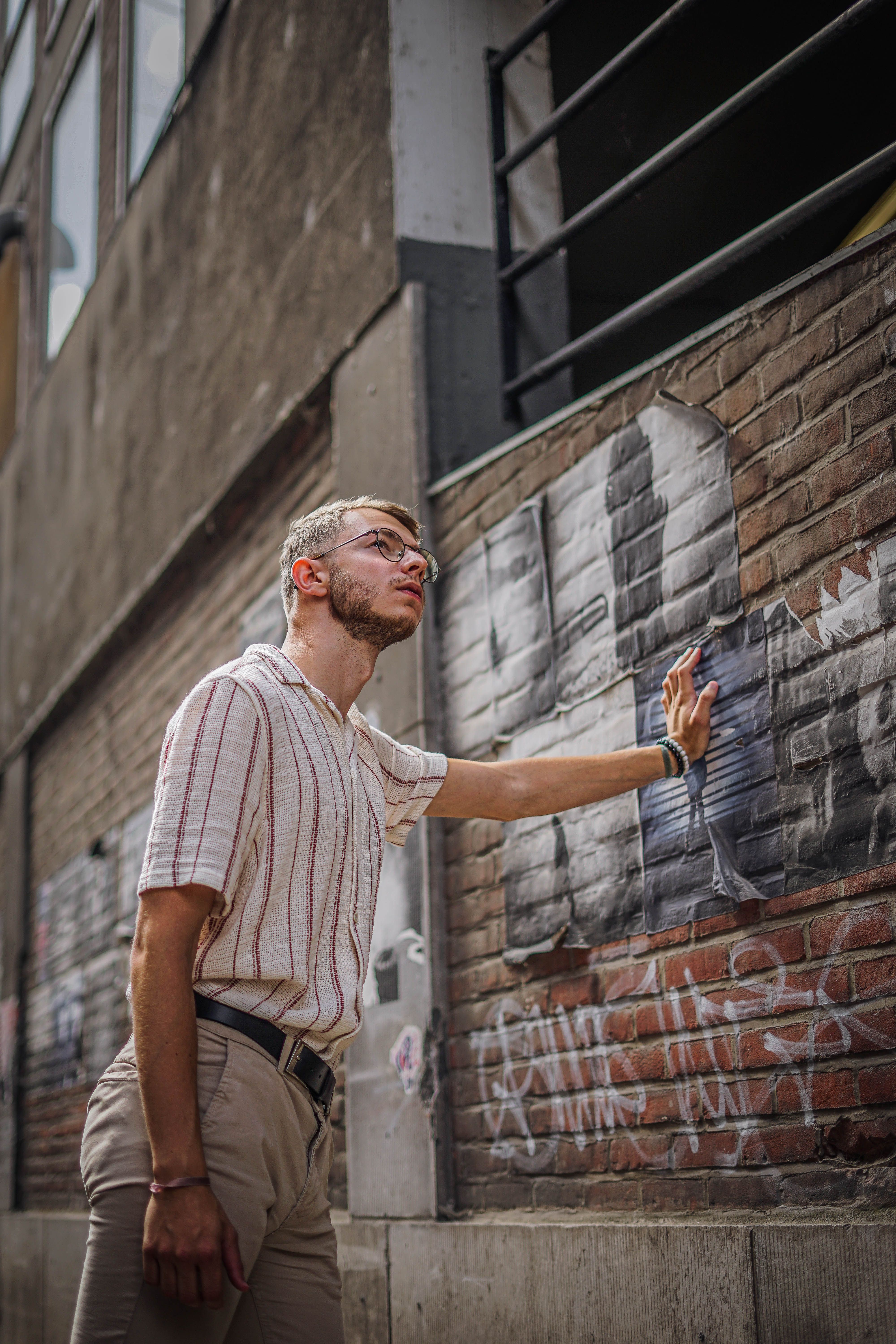 A man with glasses leans against a graffiti-covered brick wall in an urban setting.